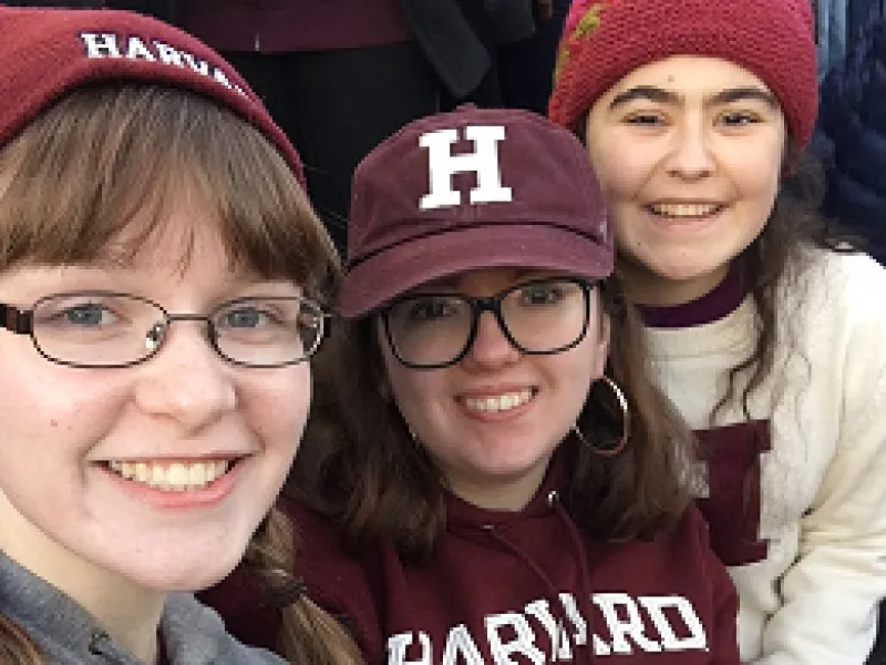 Three girls in Harvard gear take a selfie in the bleachers of a football stadium.