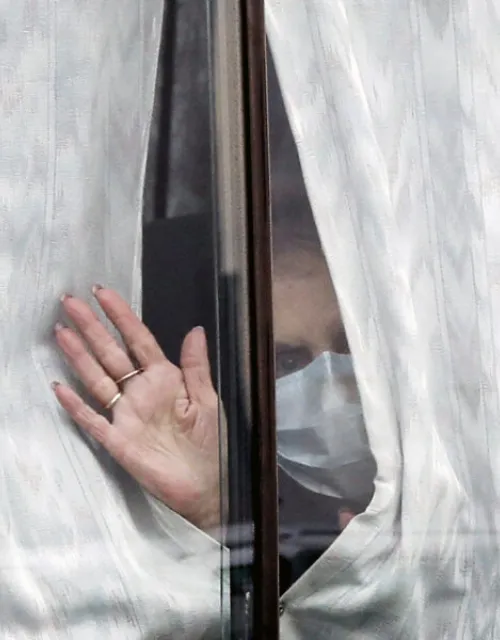 A masked woman waves hello through a window.