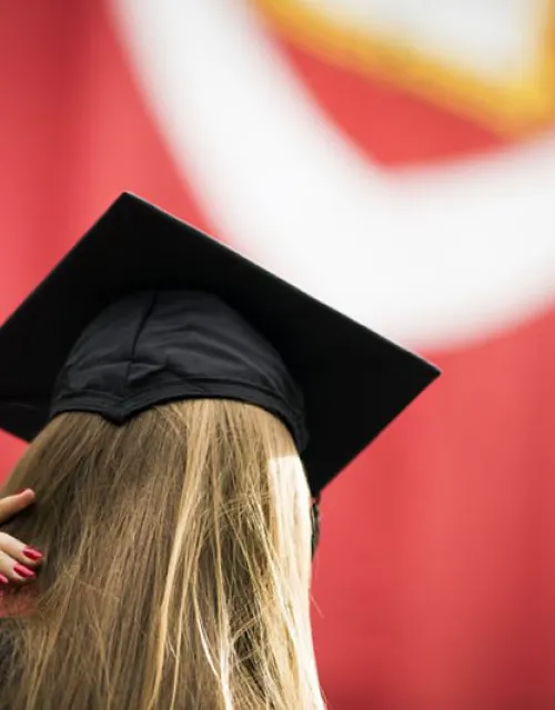back of a  student's head - shown wearing cap and gown with a banner in the background with the veritas shield