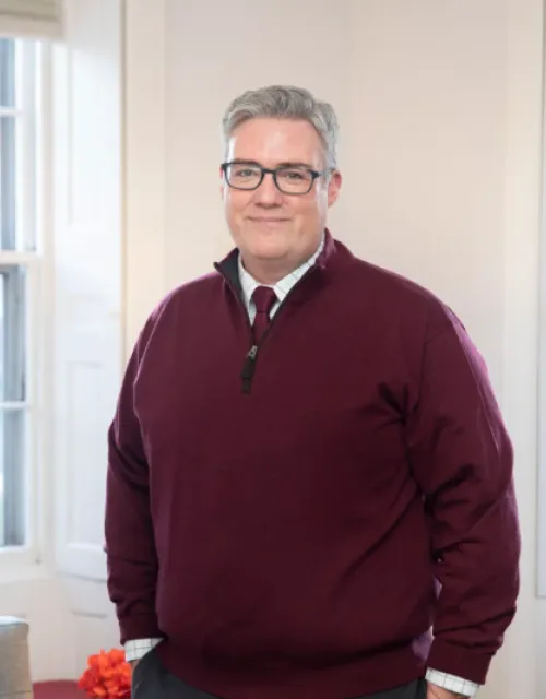Image of the new Dean of Students, Thomas Dunne, standing in front of windows in the corner of a room. 