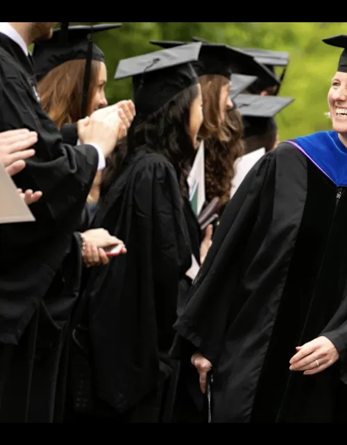 FAS dean Hopi Hoekstra, wearing a graduation cap and gown, smiles as she walks alongside an applauding group of cap- and gown-wearing graduates.