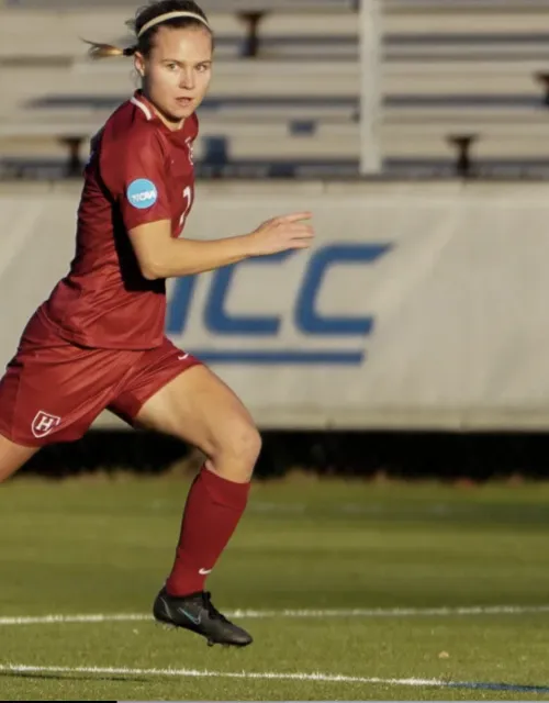 Harvard soccer player Josefine Hasbo, wearing the Harvard team's crimson uniform, dribbles the ball up the field. Some bleachers and a banner with the ACC logo lie in the background.