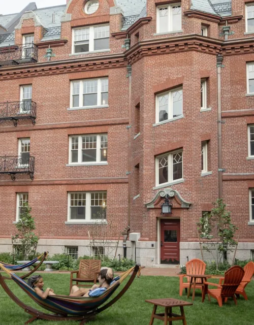 Image of a brick building with windows known as Randolph Hall, with two people sitting in a Hammock outside on a green lawn. 