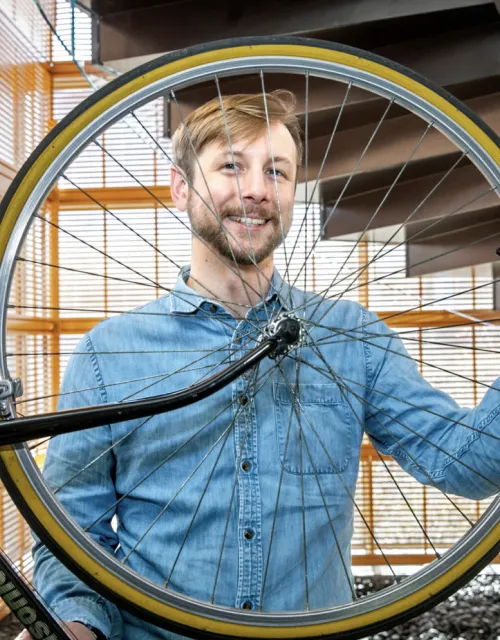 Cory Gills standing and holding up a bike wheel in front of his face. 