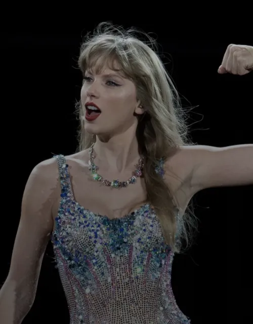 Taylor swift standing in a sparkly body suite with her left arms held up flexing her bicep. 