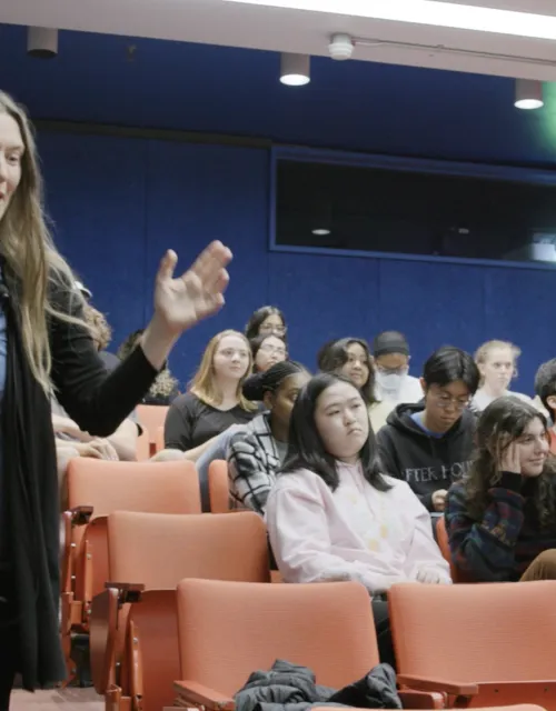female professor walking around the class  - The classroom has stadium-type seating, and the professor is holding a blue cube that says "catch box".