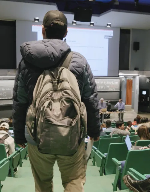 back of male student shown wearing backpack and walking into class