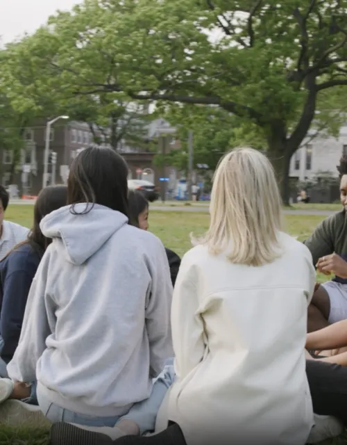 Group of students sit on banks of Charles River in circle 
