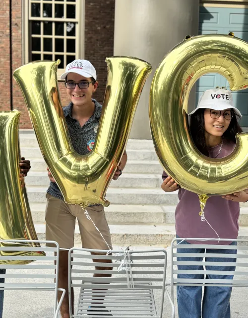 Three students holding large gold balloons forming "HVC" (Harvard Votes Challenge). They wear "VOTE" hats, situated in front of a campus building with white and red balloons nearby, smiling in a lively, outdoor setting.