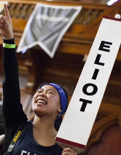 A person smiling and cheering, holding an "ELIOT" sign, with banners and a wooden interior background.