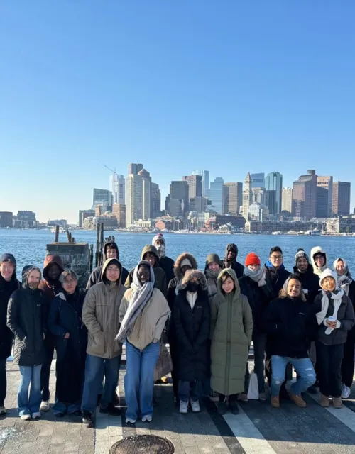 A group of people in winter clothing standing on a waterfront with a city skyline in the background under a clear blue sky.