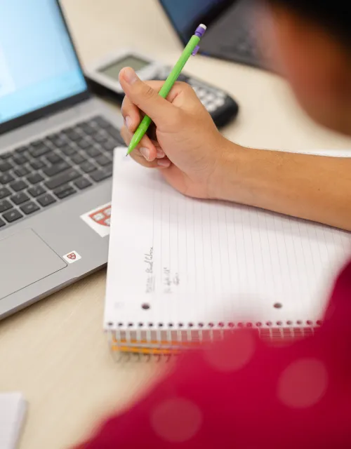 A person is taking notes in a spiral notebook with a green pencil while looking at a laptop screen