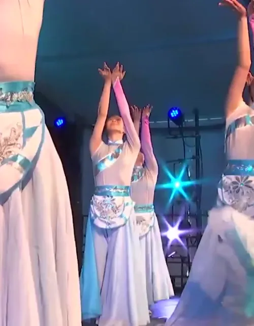 Dancers in flowing blue and white costumes performing on stage with arms raised.