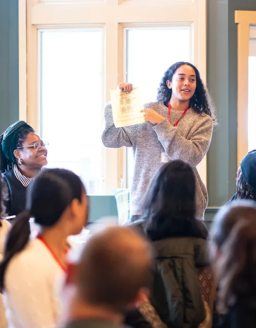 A woman stands and holds up a piece of paper while speaking to a group of people seated around her.