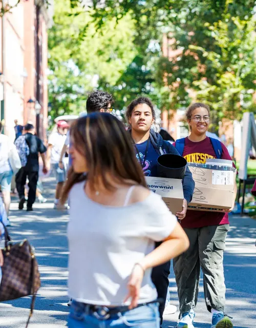 Students walk on campus, some carrying boxes and supplies, during move-in day.