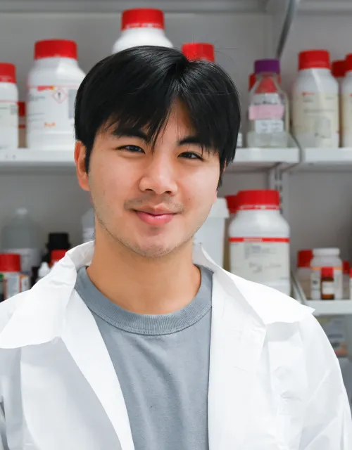 Sean stands in his white lab coat in front of a wall of medicine.