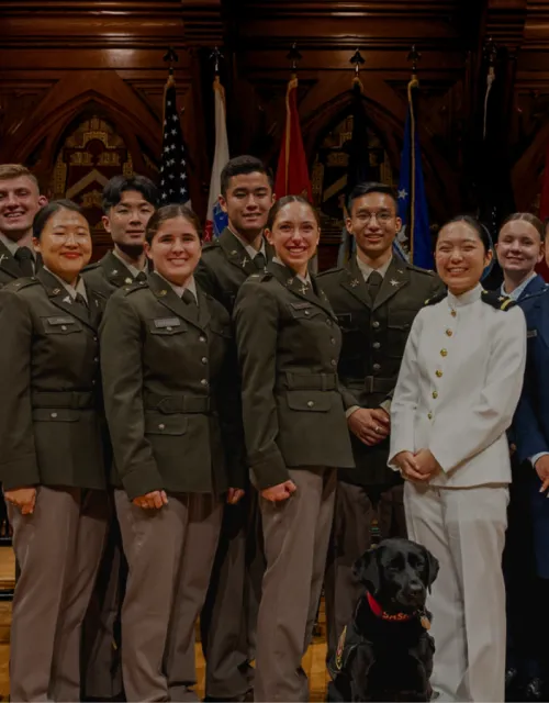 A group of ROTC cadets in uniform pose for a photo indoors with flags in the background.