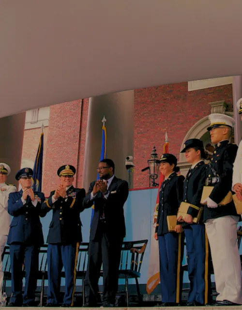 A group of military personnel and officials stand and applaud on an outdoor stage under a tent.