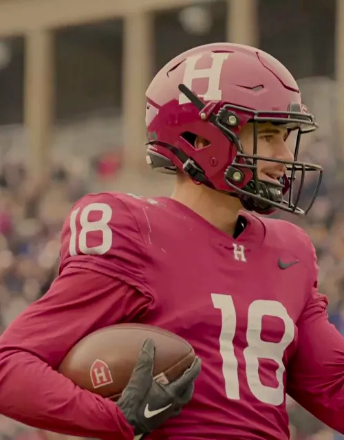 Harvard football player in a red uniform holds a football and points, with a stadium crowd in the background.