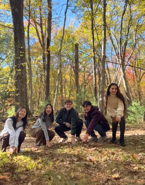 A group of students crouch and smile while touching the ground of Harvard Forest.