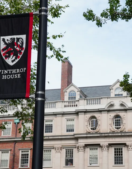 A banner with a crest and "Winthrop House" hangs on a pole in front of a historic brick and stone building, surrounded by trees.