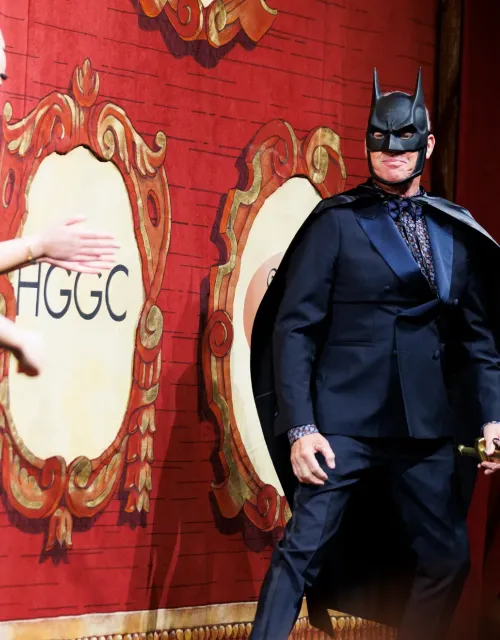 Person in a Batman mask and cape stands on stage in a suit, with a red curtain and another person clapping.