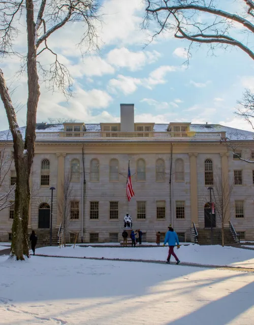 Historic campus building with an American flag and statue in front, surrounded by snow and tall trees with people walking.