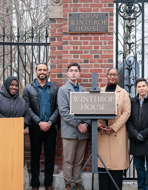 Group of people stand smiling by the Winthrop House gate and sign during an outdoor ceremony.