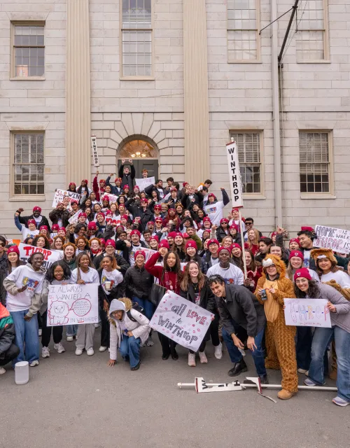 Large group of students in Winthrop gear cheer and hold signs in front of a campus building.