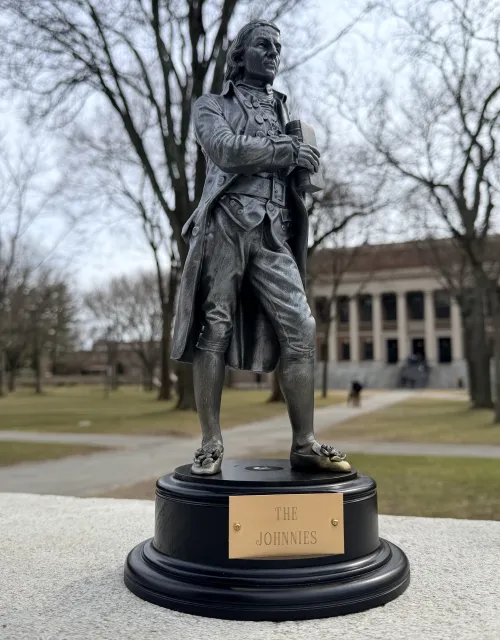 Small bronze trophy of a colonial-era man labeled “The Johnnies” sits on a ledge in a tree-lined college quad with a large library in the background.