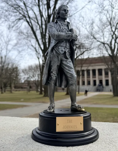Small bronze trophy of a colonial-era man labeled “The Johnnies” sits on a ledge in a tree-lined college quad with a large library in the background.