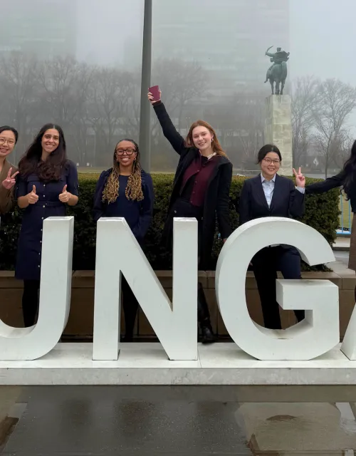 Six people pose smiling behind large “#UNGA” letters outside on a foggy day.