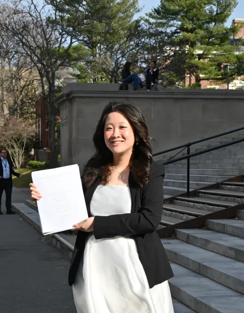 Lani Tran '26 standing on the steps of Widener library holding her thesis pages.