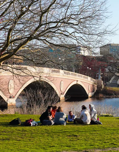 Students relax on a grassy riverbank near a stone bridge, with brick dorm buildings across the water.