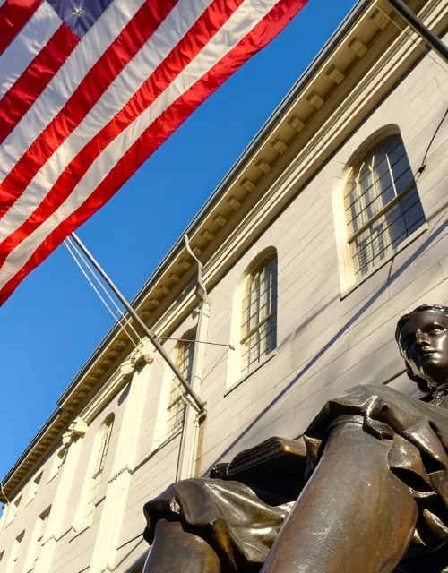 John Harvard statue outside a stone campus building beneath an American flag on a clear day.