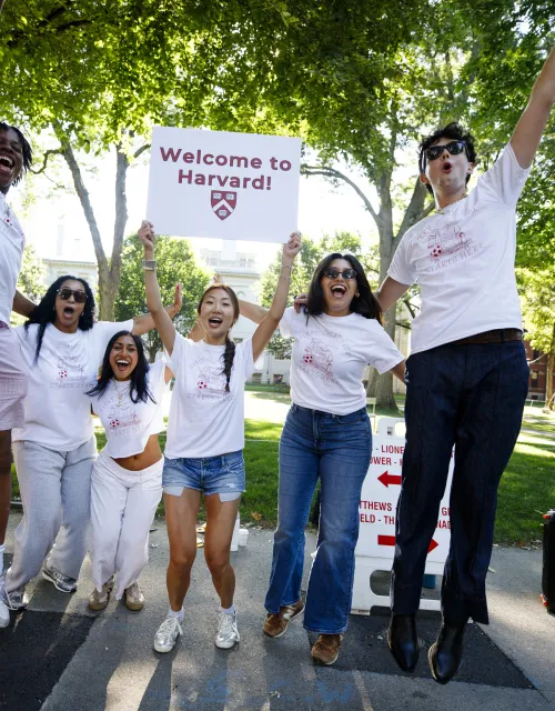 Five students jumping up in the air and cheering while holding a "welcome to harvard" sign.
