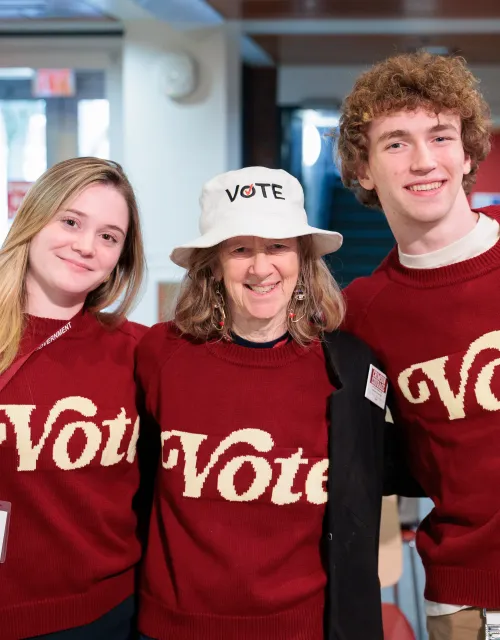 Two students (left and right) and a woman (middle) wearing matching red sweatshirts that say "Vote".