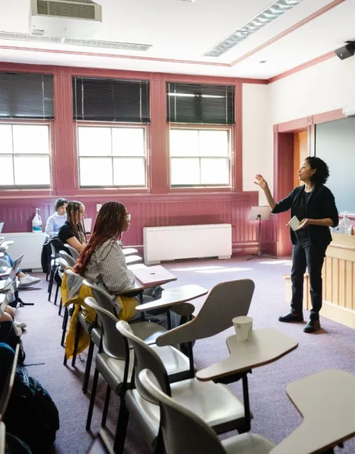 A female professor standing in the front of the classroom teaching to a room of students seated at their desks.