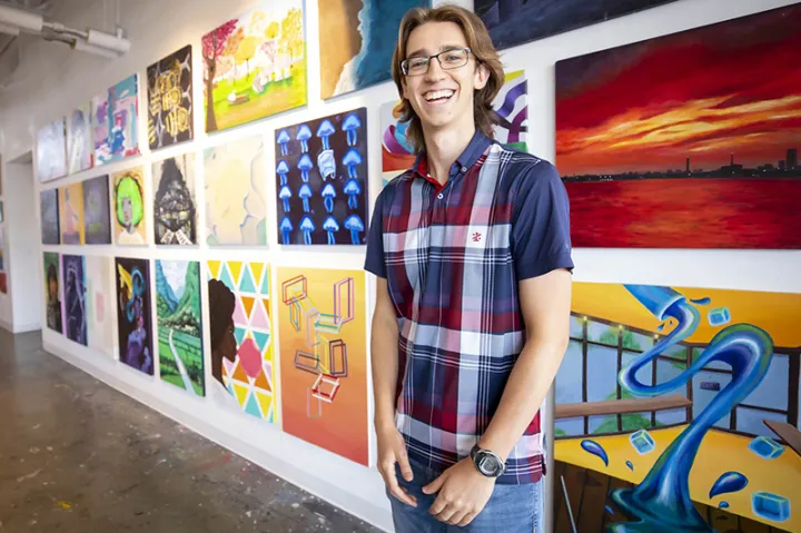 First-year student, Ezra Feder, standing in front of paintings and smiling.