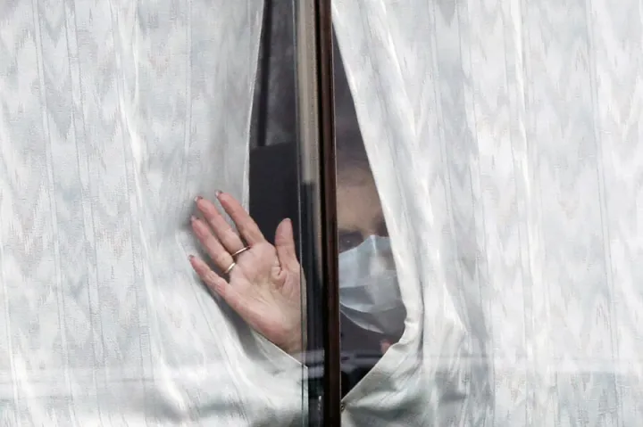 A masked woman waves hello through a window.