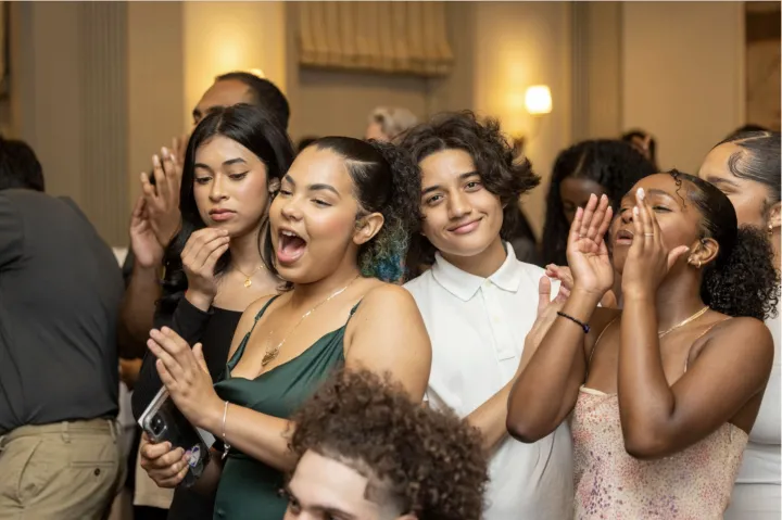 Several students dressed in formal attire dance, sing, and shout in a well-lit room. One of the students is smiling directly at the camera.