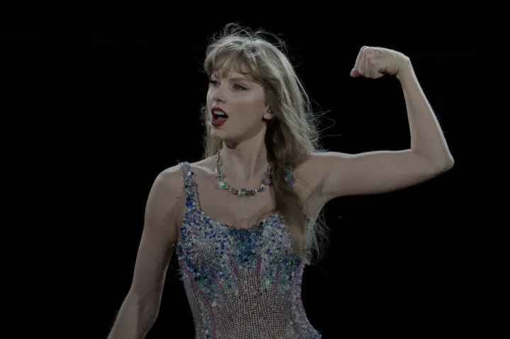 Taylor swift standing in a sparkly body suite with her left arms held up flexing her bicep. 
