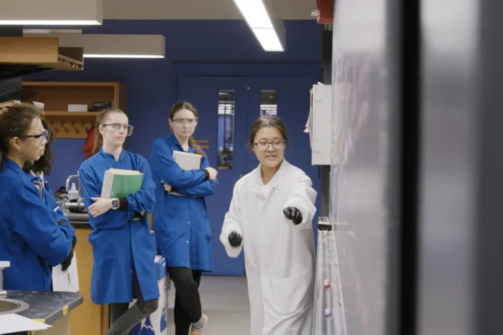 students in a lab with a professor. they are all wearing lab coats