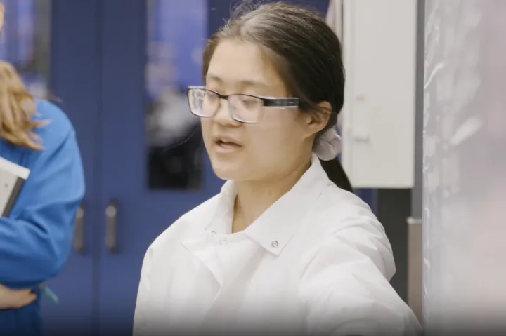 Student in lab coat and goggles at a white board