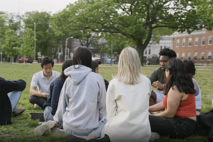 Group of students sit on banks of Charles River in circle 