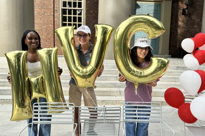 Three students holding large gold balloons forming "HVC" (Harvard Votes Challenge). They wear "VOTE" hats, situated in front of a campus building with white and red balloons nearby, smiling in a lively, outdoor setting.
