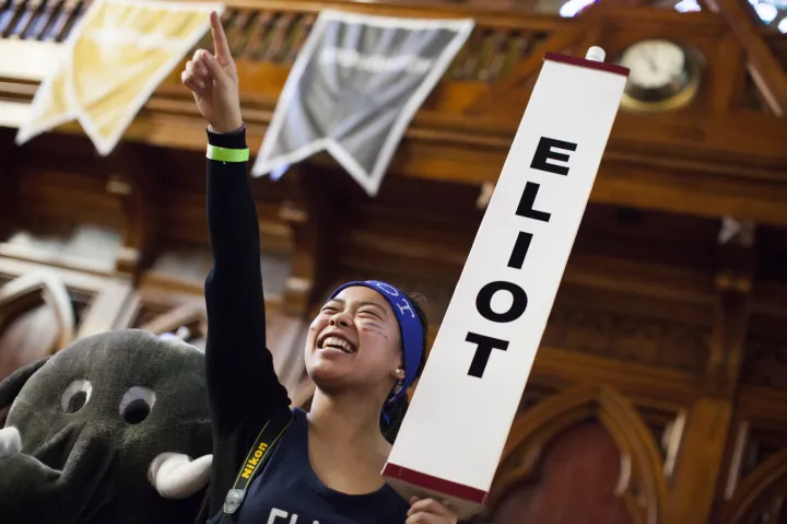 A person smiling and cheering, holding an "ELIOT" sign, with banners and a wooden interior background.