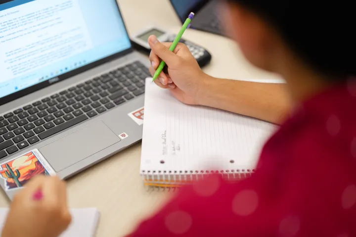 A person is taking notes in a spiral notebook with a green pencil while looking at a laptop screen