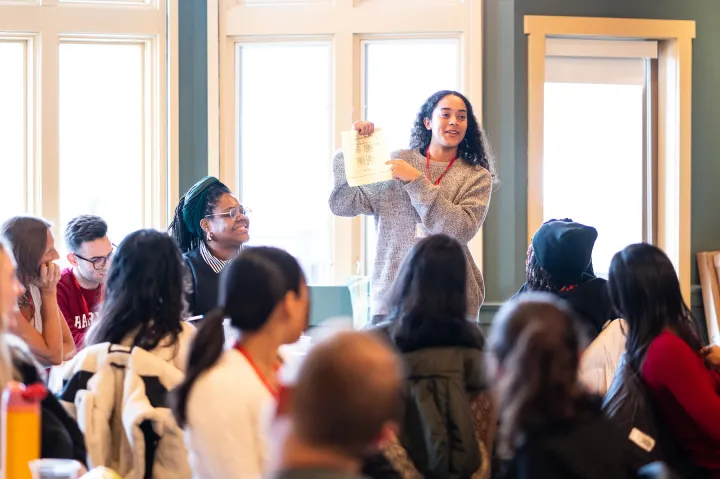 A woman stands and holds up a piece of paper while speaking to a group of people seated around her.