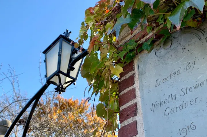A brick pillar with an engraved plaque and ivy next to a black lamp post against a blue sky.